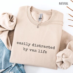 May include: A beige sweatshirt with the text "easily distracted by van life" in black. A silver necklace with a sun pendant is on the sweatshirt. The sweatshirt is folded on top of a pair of blue jeans.