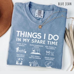 May include: Blue jean colored t-shirt with the text "THINGS I DO IN MY SPARE TIME" and gardening-related icons. A gold necklace with a small pendant rests on the shirt. The shirt is folded with a brown hat visible.