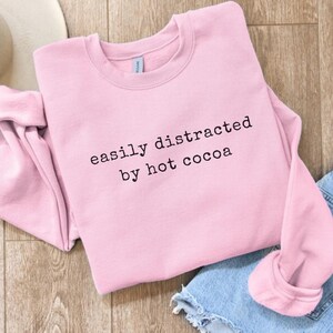May include: A light pink crewneck sweatshirt featuring the black text "easily distracted by hot cocoa" printed across the chest. The garment has long sleeves, a ribbed collar, and a soft fleece interior, presented in a casual, everyday style.