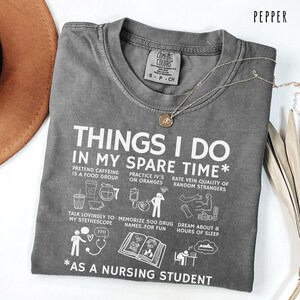 Peut inclure: T-shirt gris poivre avec le texte blanc "THINGS I DO IN MY SPARE TIME* AS A NURSING STUDENT". Le t-shirt pr&eacute;sente des illustrations d'activit&eacute;s li&eacute;es aux soins infirmiers. Un collier argent&eacute; avec un pendentif est drap&eacute; sur le t-shirt.