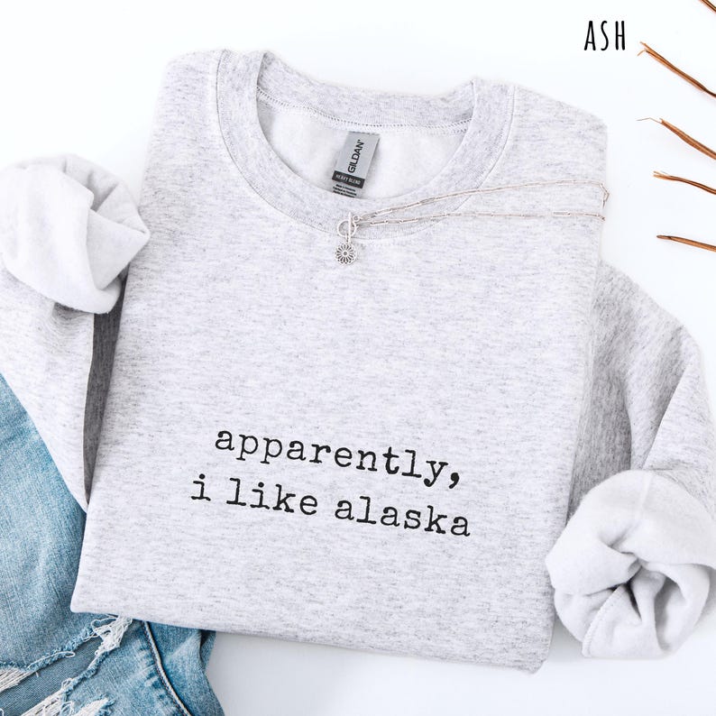 May include: A light gray sweatshirt with the text "apparently, i like alaska" in black. A silver necklace with a small pendant is draped across the front. The sweatshirt is folded on a white surface, with a pair of blue jeans visible.