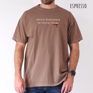 May include: Espresso-colored t-shirt with the text "easily distracted by horror films" in white, and a red blood drip graphic. The shirt has short sleeves and a crew neck.