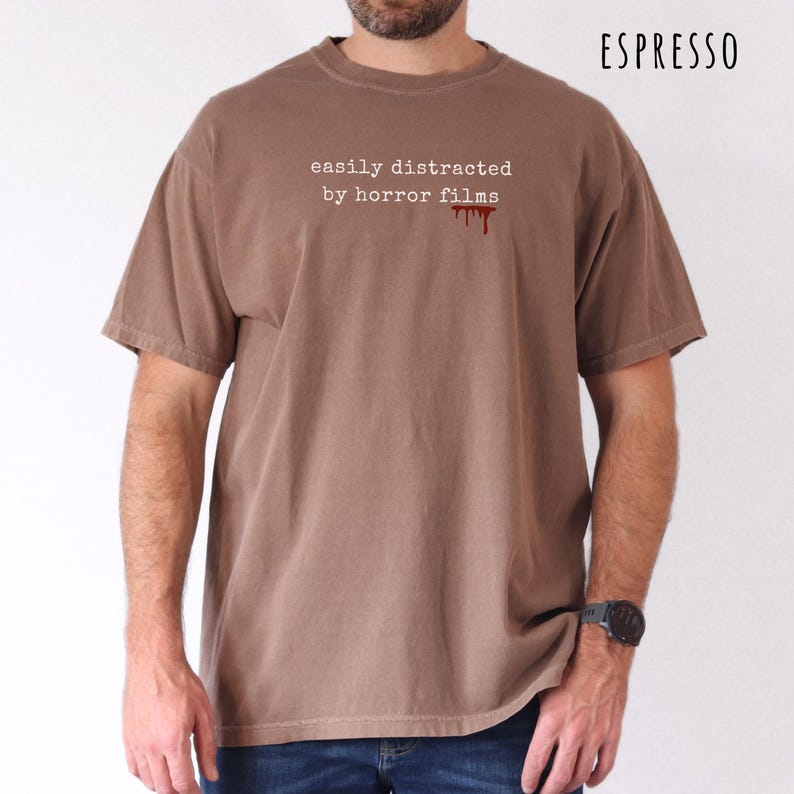 May include: Espresso-colored t-shirt with the text "easily distracted by horror films" in white, and a red blood drip graphic. The shirt has short sleeves and a crew neck.
