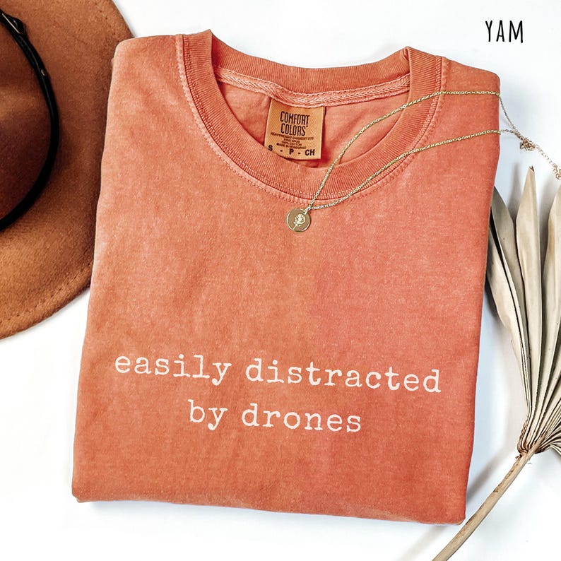 May include: A coral-colored t-shirt with the text "easily distracted by drones" in white. A gold necklace with a pendant is draped over the shirt. The shirt is folded, and a brown hat is visible in the background.