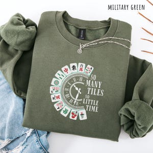 May include: Military green sweatshirt with a crew neck and long sleeves. The front features a graphic of a clock and Mahjong tiles with the text "SO MANY TILES SO LITTLE TIME". A silver necklace with a pendant is draped on the sweatshirt.