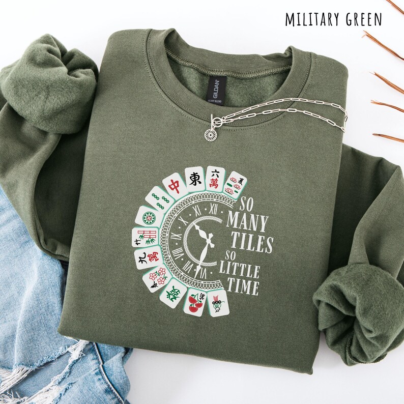 May include: Military green sweatshirt with a crew neck and long sleeves. The front features a graphic of a clock and Mahjong tiles with the text "SO MANY TILES SO LITTLE TIME". A silver necklace with a pendant is draped on the sweatshirt.