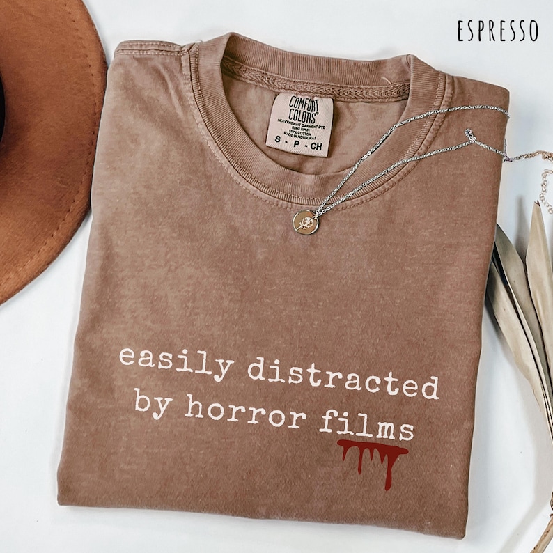 May include: Espresso-colored t-shirt with the text "easily distracted by horror films" in white. The shirt has a small blood drip graphic. A silver necklace with a pendant is draped across the shirt.