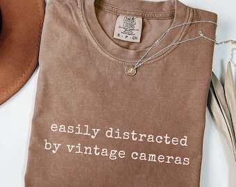 May include: Espresso-colored t-shirt with the text "easily distracted by vintage cameras" in white. A silver necklace with a small pendant rests on the shirt. The shirt has a crew neck and short sleeves.