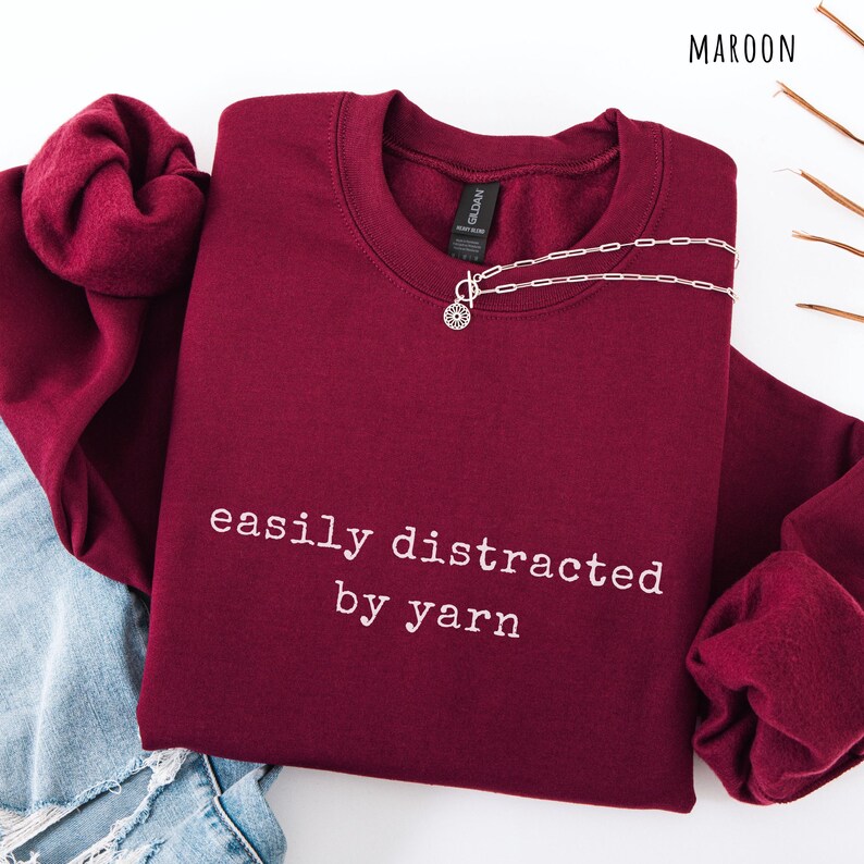 May include: Maroon crewneck sweatshirt with the text "easily distracted by yarn" in white. A silver necklace with a floral pendant is draped across the front. The sweatshirt is folded on top of a pair of blue jeans.