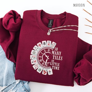 May include: Maroon sweatshirt with a circular Mahjong tile design and the text "SO MANY TILES SO LITTLE TIME." A silver necklace with a pendant is draped on the sweatshirt. The sweatshirt is folded on top of a pair of blue jeans.