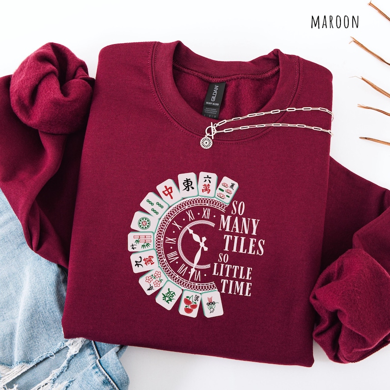 May include: Maroon sweatshirt with a circular Mahjong tile design and the text "SO MANY TILES SO LITTLE TIME." A silver necklace with a pendant is draped on the sweatshirt. The sweatshirt is folded on top of a pair of blue jeans.