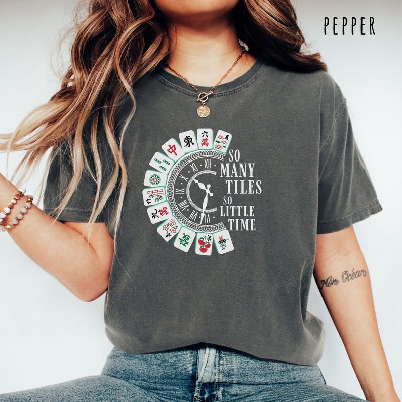 May include: A dark gray t-shirt featuring a clock design with Mahjong tiles around the perimeter. The text "SO MANY TILES SO LITTLE TIME" is printed in white. The shirt is a casual style.