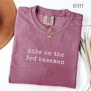 May include: Berry-colored t-shirt with the text "dibs on the 3rd baseman" in white. A gold necklace with a pendant rests on the shirt. The shirt is a Comfort Colors brand, size S-P-CH.