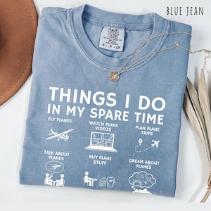 May include: Blue jean colored t-shirt with the text "THINGS I DO IN MY SPARE TIME" and illustrations of airplanes and related activities. A gold necklace with a small pendant rests on the shirt. The shirt is folded.