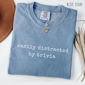 Blue jean comfort color style shirt featuring a minimalist white text design about being distracted by trivia, laid flat with a casual aesthetic. Perfect gift for trivia lovers, quiz night fans, pub quiz regulars, board game players, trivia hosts, trivia team captains, history buffs, science nerds, smart humor fans, introverts who love facts, and anyone obsessed with random knowledge and fun facts.