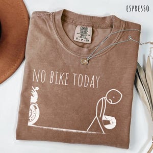 May include: Espresso-colored t-shirt with the text "NO BIKE TODAY" and a graphic of a motorcycle and a dejected stick figure. A silver necklace with a pendant is draped across the shirt.
