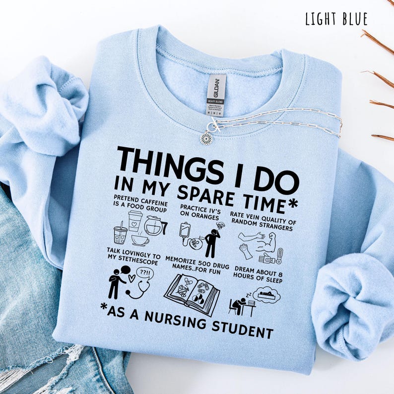Peut inclure: Sweat-shirt bleu clair avec l'inscription "THINGS I DO IN MY SPARE TIME *AS A NURSING STUDENT" et diverses illustrations en noir et blanc. Le texte comprend des phrases comme "Pretend caffeine is a food group" et "Memorize 500 drug names."
