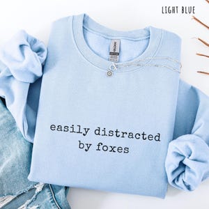 Pu&ograve; includere: Felpa azzurra con la scritta nera "easily distracted by foxes". Una collana d'argento con un ciondolo a forma di fiore &egrave; sulla felpa. La felpa &egrave; piegata sopra un paio di jeans blu.