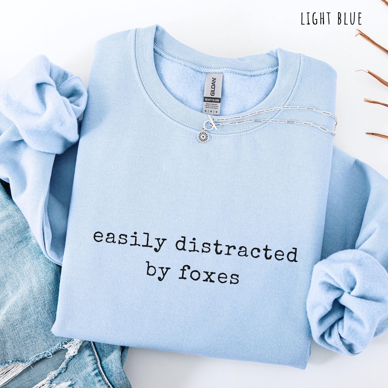 Pu&ograve; includere: Felpa azzurra con la scritta nera "easily distracted by foxes". Una collana d'argento con un ciondolo a forma di fiore &egrave; sulla felpa. La felpa &egrave; piegata sopra un paio di jeans blu.