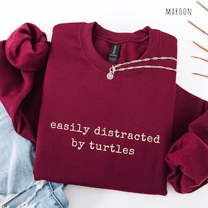 May include: A maroon sweatshirt with the text "easily distracted by turtles" in white. A silver necklace with a pendant is draped across the front. The sweatshirt is folded on a light surface.