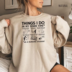 Peut inclure: Un sweat-shirt beige avec l'inscription noire "THINGS I DO IN MY SPARE TIME* AS A NURSING STUDENT". Le motif comprend des illustrations de tasses &agrave; caf&eacute;, de poches IV et d'un st&eacute;thoscope. Le mot "NATURAL" est imprim&eacute; en haut &agrave; droite.