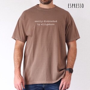 May include: Espresso-colored t-shirt with the text "easily distracted by alligators" in a simple, white font. The shirt has a classic crew neck and short sleeves. The model is wearing a black watch.