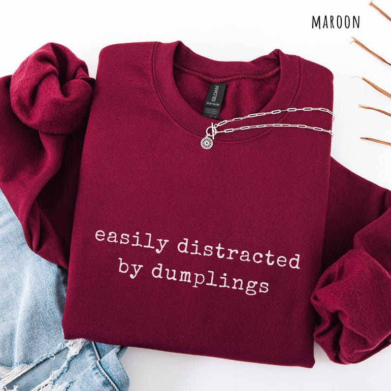 May include: Maroon sweatshirt with the text "easily distracted by dumplings" in white. A silver necklace with a circular pendant is draped across the front. The sweatshirt is folded on top of a pair of blue jeans.