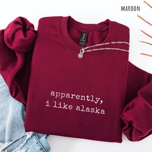 May include: Maroon crewneck sweatshirt with the text "apparently, i like alaska" in white. A silver necklace with a circular pendant is draped on the sweatshirt. The word "MAROON" is in the top right corner.