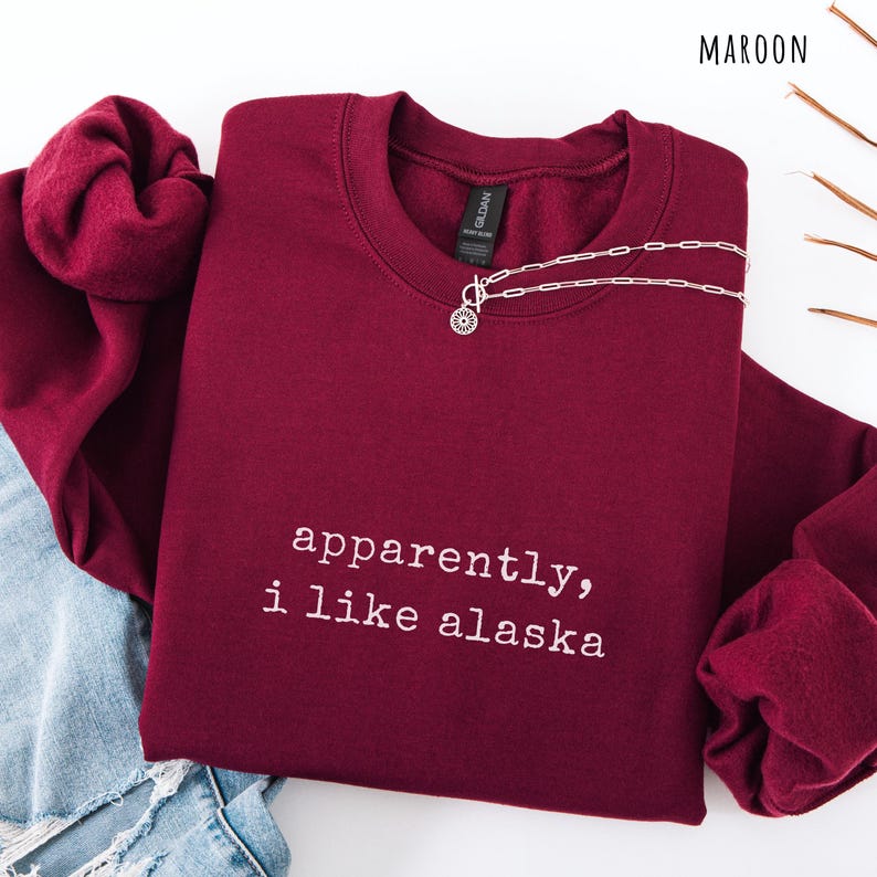 May include: Maroon crewneck sweatshirt with the text "apparently, i like alaska" in white. A silver necklace with a circular pendant is draped on the sweatshirt. The word "MAROON" is in the top right corner.