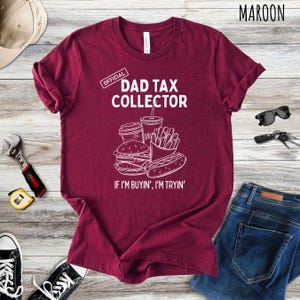 Może przedstawiać: Bordowa koszulka z napisem "DAD TAX COLLECTOR" i grafiką przedstawiającą jedzenie typu fast food. Poniżej grafiki znajduje się napis "IF I'M BUYIN', I'M TRYIN'". Koszulka to element odzieży codziennej.