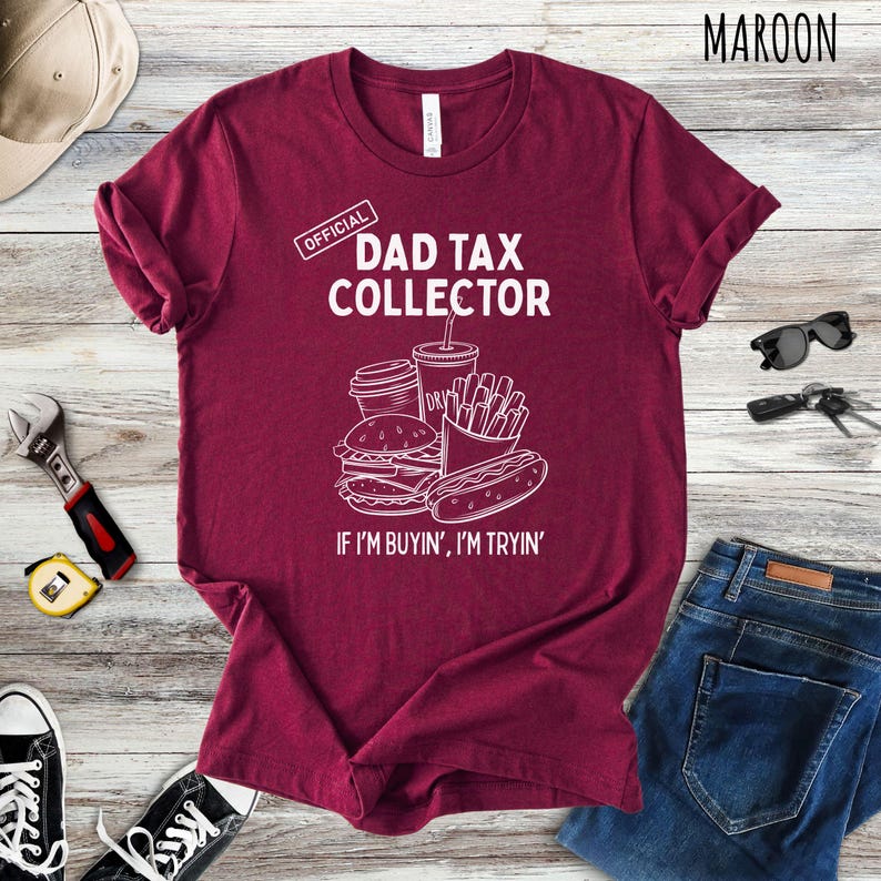 Może przedstawiać: Bordowa koszulka z napisem "DAD TAX COLLECTOR" i grafiką przedstawiającą jedzenie typu fast food. Poniżej grafiki znajduje się napis "IF I'M BUYIN', I'M TRYIN'". Koszulka to element odzieży codziennej.