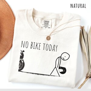May include: Cream-colored t-shirt with the text "NO BIKE TODAY" in black. A stick figure with a motorcycle helmet is depicted next to a motorcycle. A gold necklace with a pendant is draped on the shirt.