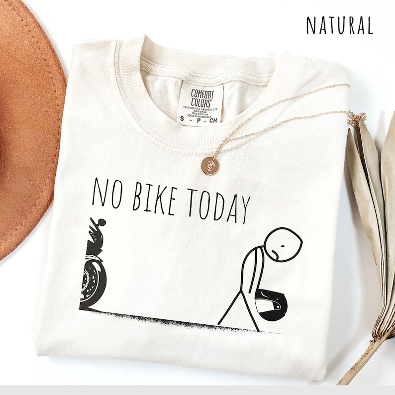 May include: Cream-colored t-shirt with the text "NO BIKE TODAY" in black. A stick figure with a motorcycle helmet is depicted next to a motorcycle. A gold necklace with a pendant is draped on the shirt.