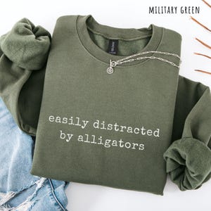 May include: Military green sweatshirt with the text "easily distracted by alligators" in white. A silver necklace with a circular pendant is on the sweatshirt. The sweatshirt is folded on top of blue jeans.