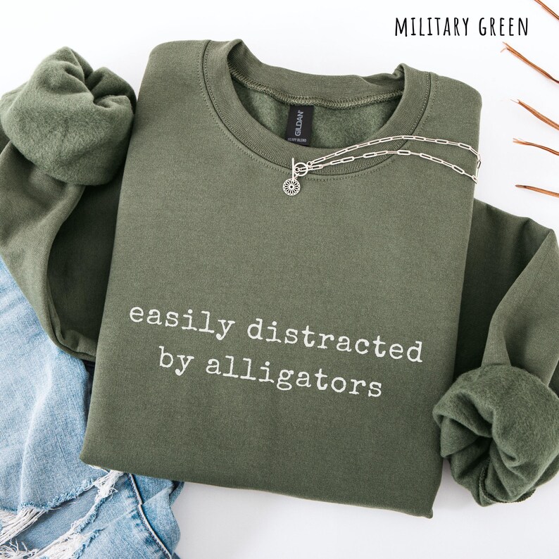 May include: Military green sweatshirt with the text "easily distracted by alligators" in white. A silver necklace with a circular pendant is on the sweatshirt. The sweatshirt is folded on top of blue jeans.