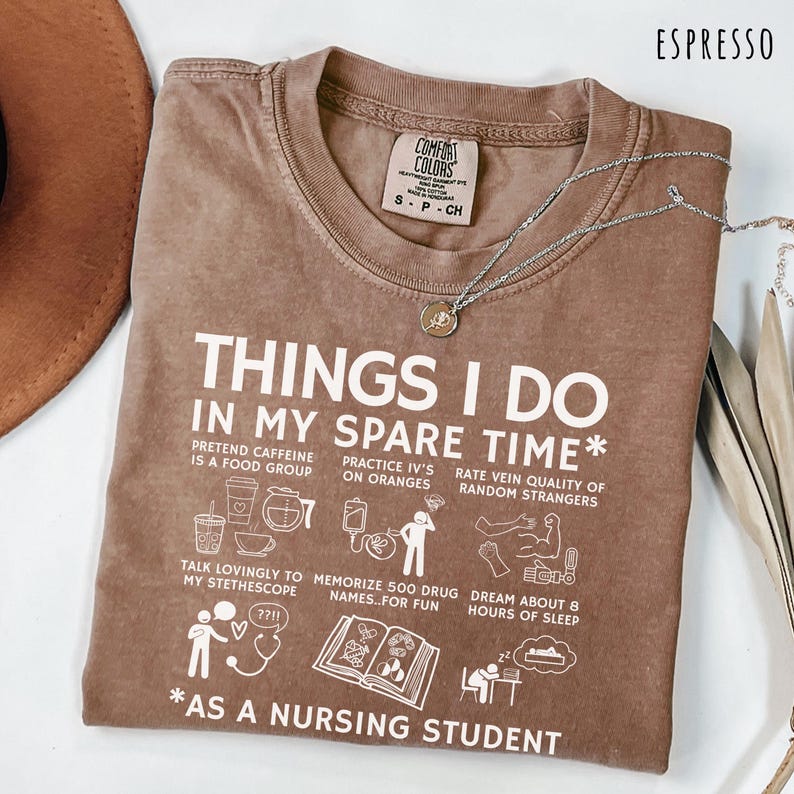 Peut inclure: T-shirt couleur expresso avec le texte "THINGS I DO IN MY SPARE TIME *AS A NURSING STUDENT" en blanc. Le t-shirt pr&eacute;sente des illustrations de tasses &agrave; caf&eacute;, de poches IV et d'un st&eacute;thoscope. Un collier argent&eacute; avec un pendentif est drap&eacute; sur le t-shirt.