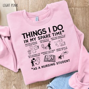 Peut inclure: Sweat-shirt rose clair avec l'inscription "THINGS I DO IN MY SPARE TIME" et des illustrations humoristiques sur la vie &eacute;tudiante en soins infirmiers. Le texte comprend des phrases comme "Pr&eacute;tendre que la caf&eacute;ine est un groupe alimentaire".