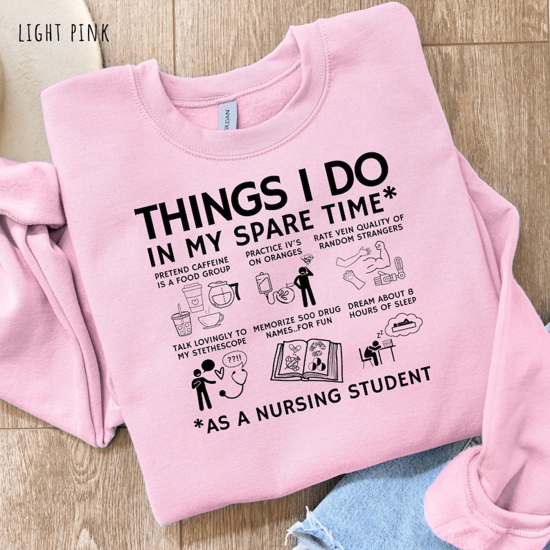 Peut inclure: Sweat-shirt rose clair avec l'inscription "THINGS I DO IN MY SPARE TIME" et des illustrations humoristiques sur la vie &eacute;tudiante en soins infirmiers. Le texte comprend des phrases comme "Pr&eacute;tendre que la caf&eacute;ine est un groupe alimentaire".