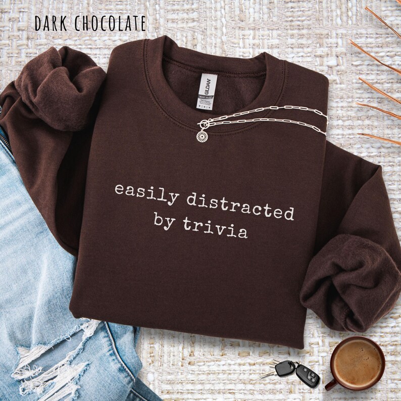 May include: Dark chocolate-colored sweatshirt with the text "easily distracted by trivia" in white. A silver necklace with a pendant is on the sweatshirt. The sweatshirt is folded on top of blue jeans. A cup of coffee and keys are also visible.