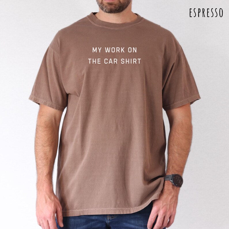 May include: Espresso-colored t-shirt with the text "MY WORK ON THE CAR SHIRT" in white. The shirt is a short-sleeved, crew neck style. The model is wearing a black watch. The shirt is a solid color.