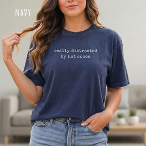 May include: A navy blue short-sleeved t-shirt featuring the white text "easily distracted by hot cocoa" printed in a typewriter-style font across the chest. The garment has a crew neckline and a relaxed fit, showcasing a solid dark blue color.