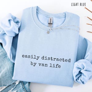 May include: Light blue crewneck sweatshirt with the text "easily distracted by van life" in black. A silver necklace with a flower pendant rests on the shirt. The sweatshirt is folded on top of a pair of blue jeans.
