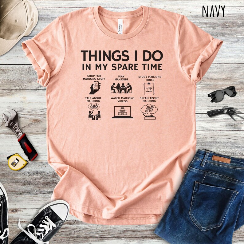 May include: A peach-colored t-shirt with the text "THINGS I DO IN MY SPARE TIME" and illustrations of mahjong-related activities. The shirt is paired with blue jeans and accessories.