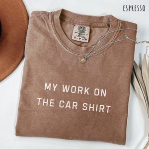 May include: Espresso-colored t-shirt with the text "MY WORK ON THE CAR SHIRT" in white block letters. The shirt has a crew neck and a small tag on the upper back. A silver necklace with a round pendant is draped across the shirt.