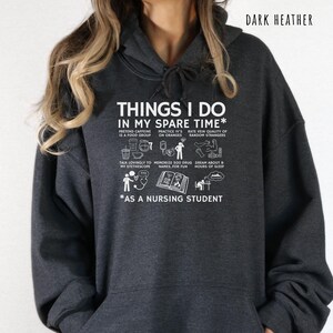 Peut inclure: Un sweat &agrave; capuche gris fonc&eacute; avec du texte blanc : "THINGS I DO IN MY SPARE TIME* AS A NURSING STUDENT". Le texte est accompagn&eacute; de petites illustrations repr&eacute;sentant diverses activit&eacute;s li&eacute;es aux soins infirmiers.