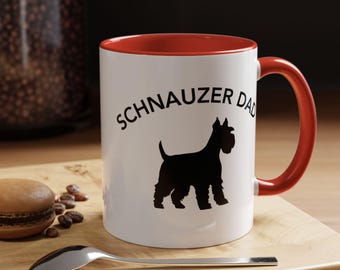 May include: White ceramic mug with a red interior and handle. The mug features the text "SCHNAUZER DAD" in black, with a black silhouette of a Schnauzer dog. The mug is on a wooden surface with a spoon and a macaron.