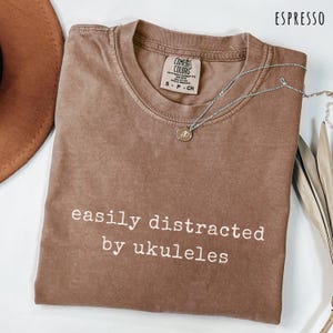 Puede incluir: Camiseta color espresso con el texto blanco "easily distracted by ukuleles". Un collar plateado con un pequeño colgante descansa sobre la camiseta. La camiseta es de Comfort Colors.