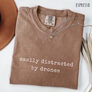 May include: Espresso-colored t-shirt with the text "easily distracted by drones" in white. The shirt has a crew neck and short sleeves. A silver necklace with a small pendant is draped across the shirt.