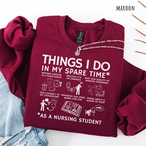 Peut inclure: Sweat-shirt bordeaux avec texte blanc : "THINGS I DO IN MY SPARE TIME" et activit&eacute;s amusantes d'&eacute;tudiant en soins infirmiers. Comprend un collier argent&eacute;. Le sweat-shirt est pli&eacute; sur un jean bleu.
