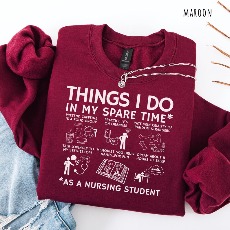Peut inclure: Sweat-shirt bordeaux avec texte blanc : "THINGS I DO IN MY SPARE TIME" et activit&eacute;s amusantes d'&eacute;tudiant en soins infirmiers. Comprend un collier argent&eacute;. Le sweat-shirt est pli&eacute; sur un jean bleu.